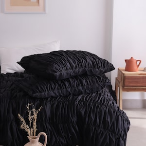 May include: A bed is made with a black ruched duvet cover and two matching pillows. A white pillow is visible. A beige vase with dried plants sits in the foreground. A framed print and a small wooden table with a terracotta teapot are also visible.