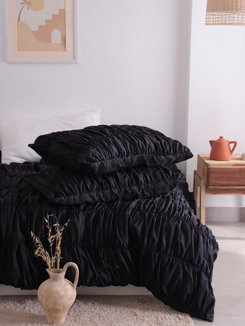 May include: A bed is made with a black ruched duvet cover and two matching pillows. A white pillow is visible. A beige vase with dried plants sits in the foreground. A framed print and a small wooden table with a terracotta teapot are also visible.