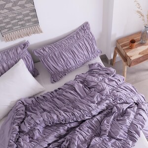 Puede incluir: Una cama est&aacute; hecha con un edred&oacute;n fruncido de color lavanda y fundas de almohada a juego. Tambi&eacute;n hay una almohada blanca en la cama. Una peque&ntilde;a mesita de noche de madera con una taza marr&oacute;n y una botella de vidrio est&aacute; al lado de la cama. Un tapiz tejido est&aacute; encima de la cama.