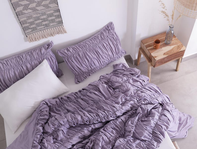 Puede incluir: Una cama est&aacute; hecha con un edred&oacute;n fruncido de color lavanda y fundas de almohada a juego. Tambi&eacute;n hay una almohada blanca en la cama. Una peque&ntilde;a mesita de noche de madera con una taza marr&oacute;n y una botella de vidrio est&aacute; al lado de la cama. Un tapiz tejido est&aacute; encima de la cama.