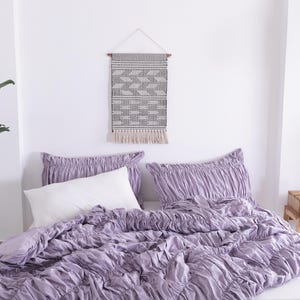 Puede incluir: Una escena de dormitorio con una cama con un edred&oacute;n lavanda y fundas de almohada con volantes a juego. Una almohada blanca est&aacute; en la cama. Un tapiz de pared con un patr&oacute;n geom&eacute;trico cuelga sobre la cama. Una planta en maceta y una mesita de noche tambi&eacute;n est&aacute;n en la habitaci&oacute;n.
