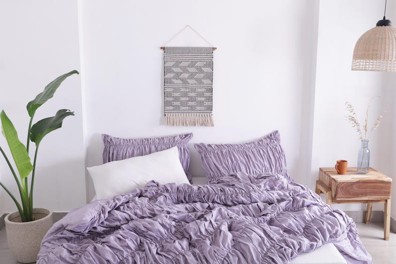 Puede incluir: Una escena de dormitorio con una cama con un edred&oacute;n lavanda y fundas de almohada con volantes a juego. Una almohada blanca est&aacute; en la cama. Un tapiz de pared con un patr&oacute;n geom&eacute;trico cuelga sobre la cama. Una planta en maceta y una mesita de noche tambi&eacute;n est&aacute;n en la habitaci&oacute;n.