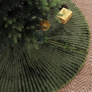 Puede incluir: Una falda de &aacute;rbol de Navidad de terciopelo verde con un dise&ntilde;o plisado. La falda circular tiene un borde verde. Se ve un &aacute;rbol de Navidad con adornos dorados y una caja de regalo envuelta.