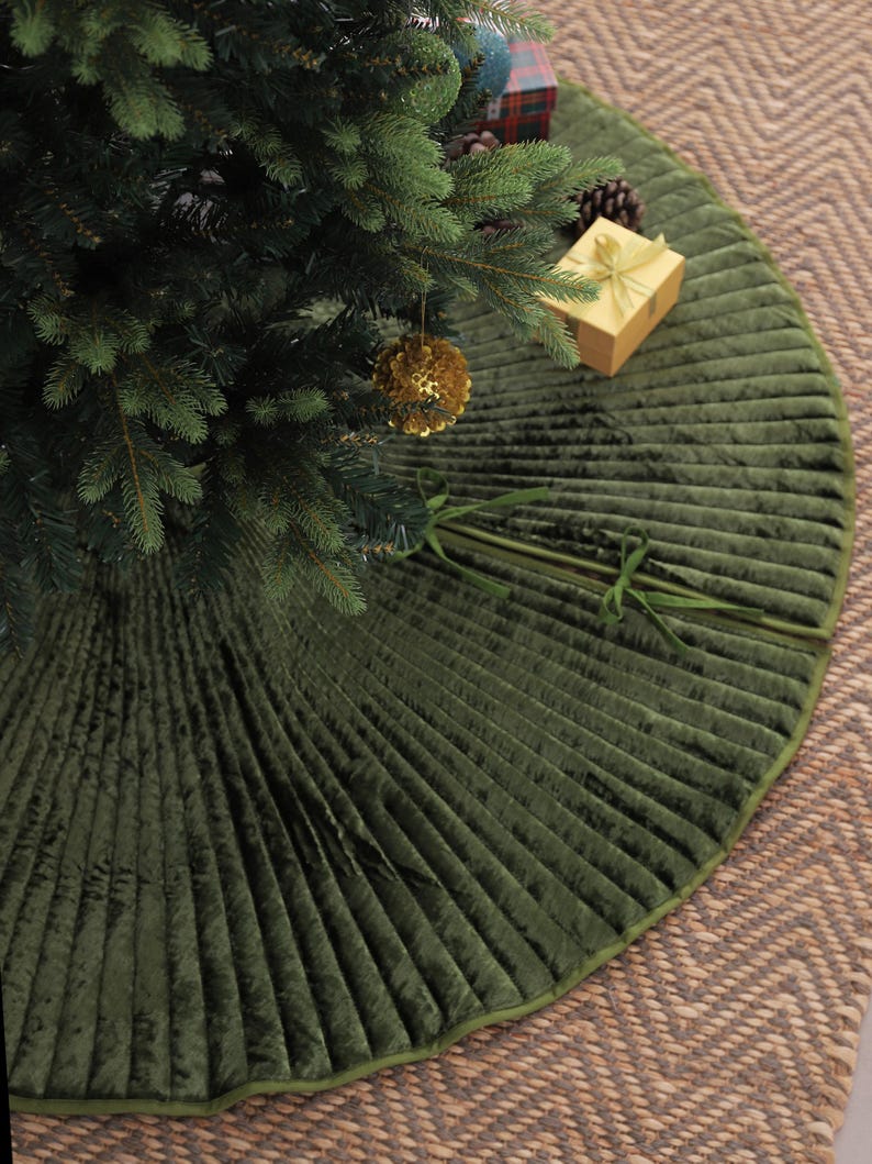 Puede incluir: Una falda de &aacute;rbol de Navidad de terciopelo verde con un dise&ntilde;o plisado. La falda circular tiene un borde verde. Se ve un &aacute;rbol de Navidad con adornos dorados y una caja de regalo envuelta.