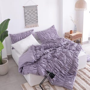 Puede incluir: Una cama con un edred&oacute;n fruncido lavanda y fundas de almohada a juego. Una almohada blanca est&aacute; en la cama. Una l&aacute;mpara de escritorio negra est&aacute; sobre un libro. Un tapiz de pared tejido y una planta en maceta tambi&eacute;n est&aacute;n en la habitaci&oacute;n.