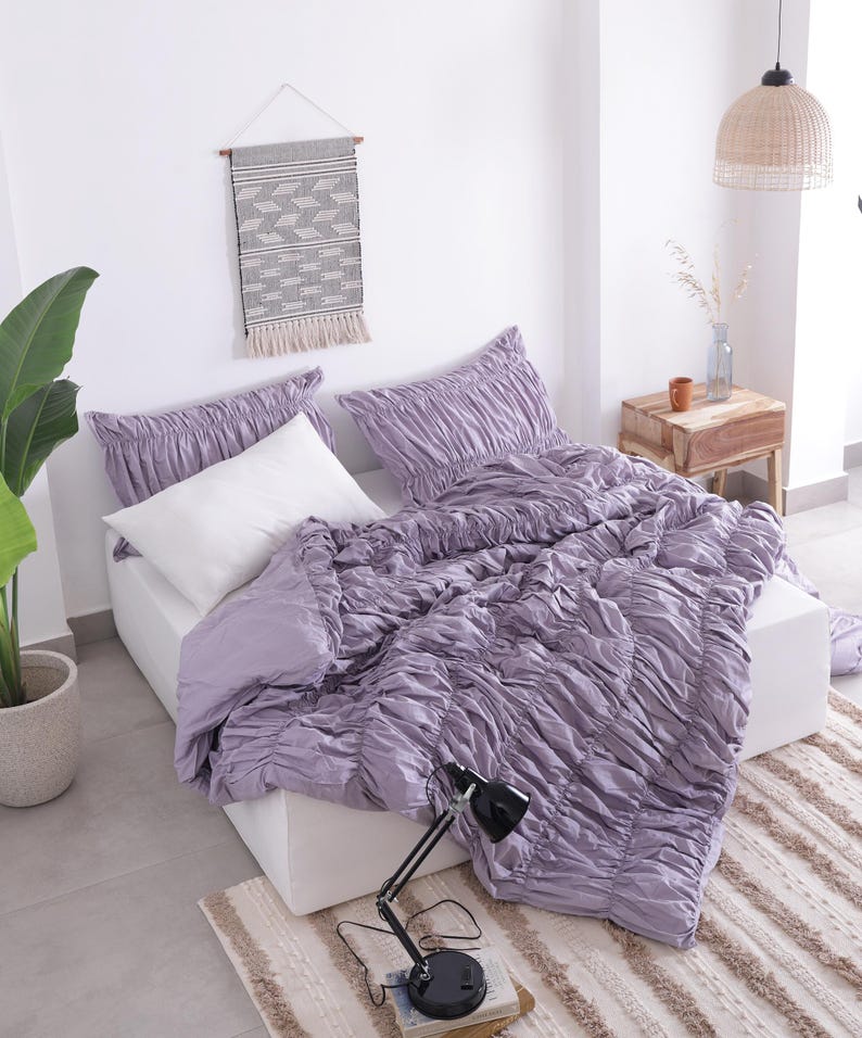 Puede incluir: Una cama con un edred&oacute;n fruncido lavanda y fundas de almohada a juego. Una almohada blanca est&aacute; en la cama. Una l&aacute;mpara de escritorio negra est&aacute; sobre un libro. Un tapiz de pared tejido y una planta en maceta tambi&eacute;n est&aacute;n en la habitaci&oacute;n.