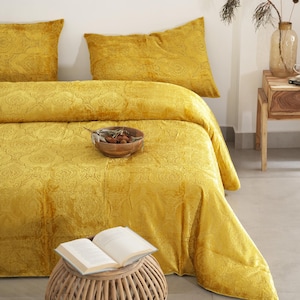 May include: A bed dressed with a mustard yellow paisley patterned duvet cover and matching pillowcases. A small wooden bowl with nuts sits on the duvet. An open book rests on a small woven side table.