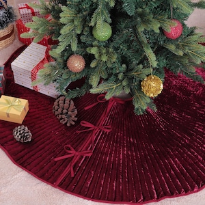 May include: A Christmas tree skirt in a deep red, pleated velvet. The skirt is circular with a ribbon trim and three decorative bows. The tree is decorated with ornaments and sits on a light-colored rug. Gift boxes and a basket are nearby.