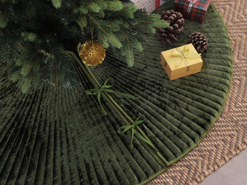 Puede incluir: Una falda de &aacute;rbol de Navidad de terciopelo plisado verde oscuro con un borde verde a juego. La falda est&aacute; decorada con un adorno dorado, una peque&ntilde;a caja de regalo y pi&ntilde;as. La falda tiene aproximadamente 127 cm de di&aacute;metro.