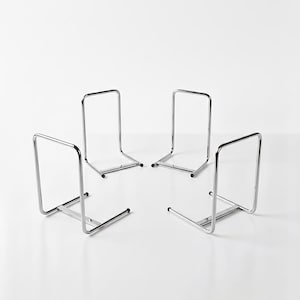 Puede incluir: Cuatro sujetalibros de metal cromado. Cada sujetalibros tiene un marco rectangular con una base horizontal y un soporte vertical. Los sujetalibros están dispuestos en un patrón circular sobre una superficie blanca.