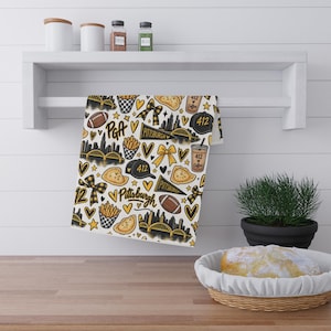 May include: A white kitchen towel featuring a repeating pattern of Pittsburgh-themed illustrations in black and yellow, including footballs, city skylines, bridges, pierogies, chips, bows, and cups labelled 412 Coffee and 412.