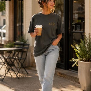 Peut inclure: Un t-shirt gris anthracite à manches courtes avec le nombre 412 brodé en fil doré sur la poitrine gauche, associé à un jean bleu délavé et des baskets blanches. Le t-shirt présente une encolure ronde et une coupe décontractée et ample.