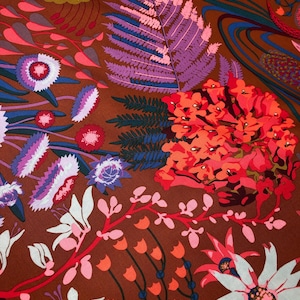 May include: A close-up of a fabric with a floral pattern. The design features large flowers in white, pink, and orange, along with purple fern-like leaves and blue accents, all set against a brown background.