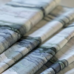 May include: Close-up of folded fabric with a watercolor-like design in shades of blue, gray, and green. The fabric has a soft, flowing appearance, with visible folds and textures. The pattern resembles abstract landscapes or flowing water.