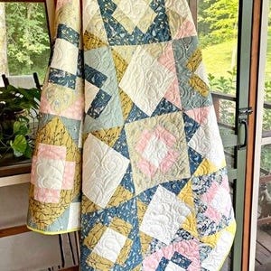 A colorful quilt with a geometric pattern hangs on a wooden ladder, with a view of a lush green outdoor area through a glass door.