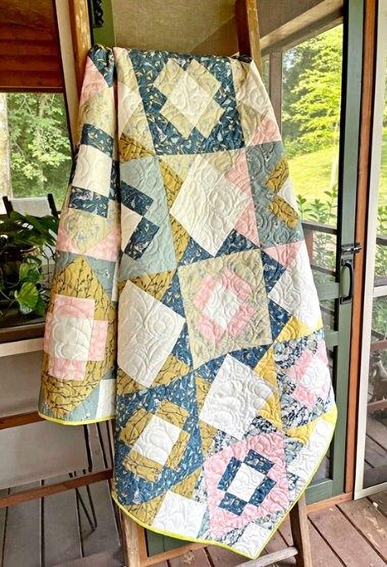 A colorful quilt with a geometric pattern hangs on a wooden ladder, with a view of a lush green outdoor area through a glass door.
