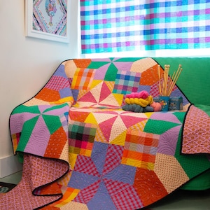May include: A colorful patchwork quilt draped over a green sofa. The quilt features geometric patterns in shades of orange, yellow, purple, and blue. Knitting needles and yarn balls are on the sofa. A framed artwork and a plaid curtain are in the background.