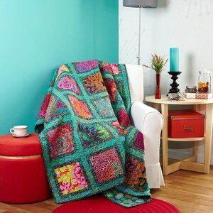 a vibrant and colorful quilt draped over a white armchair, with a red ottoman and a small table nearby. The room has a teal accent wall, and there are various decorative items such as a vase, a candle, and a jar on the table.