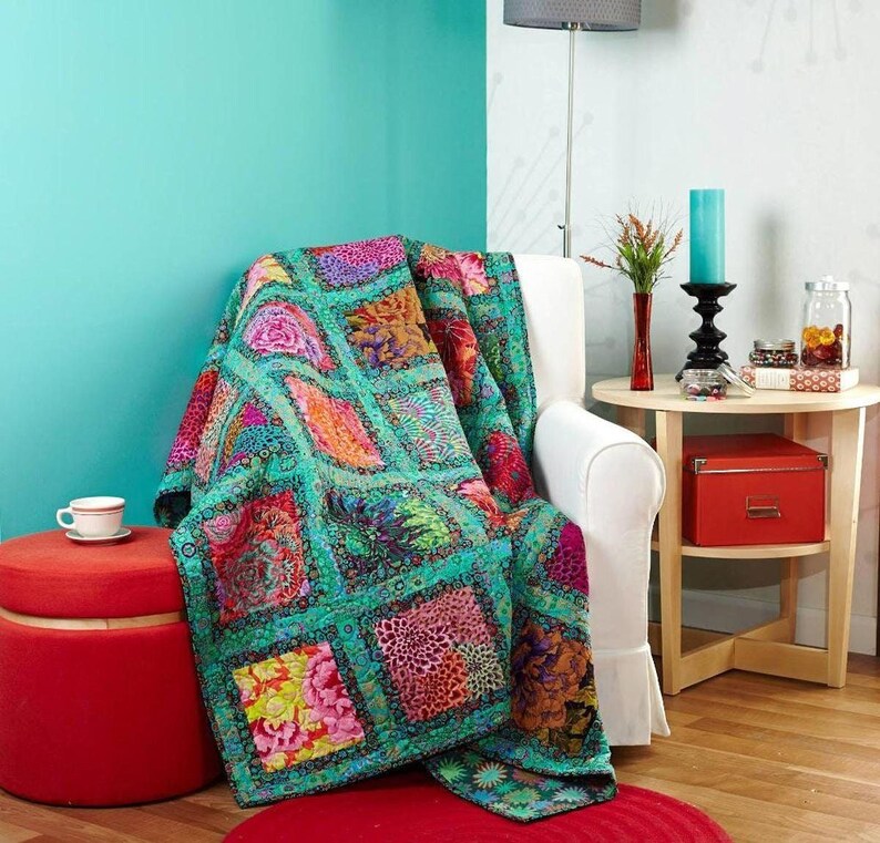 a vibrant and colorful quilt draped over a white armchair, with a red ottoman and a small table nearby. The room has a teal accent wall, and there are various decorative items such as a vase, a candle, and a jar on the table.
