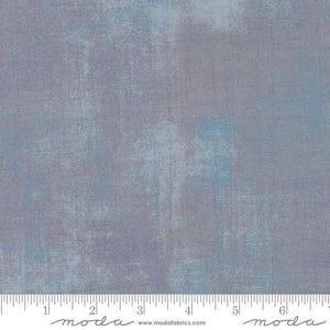 May include: A close-up of a light blue and grey fabric with a subtle textured pattern. The fabric is a solid colour with a slight variation in the shade of blue. The fabric is measured in inches with a ruler that says "moda" on it.