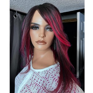May include: A mannequin head with long, dark hair and vibrant red highlights. The mannequin is wearing a white, open-knit sweater over a pink top. The face has detailed makeup, including dark eye shadow and a neutral lip color.