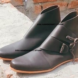 May include: A pair of dark brown leather ankle boots with a low heel and a rounded toe. One boot features a strap with a buckle and a decorative tie. The text "USA HANDICRAFT STORE" is printed on the side of each boot.