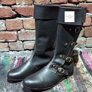 Puede incluir: Un par de botas de cuero marrón oscuro con la parte superior plegada. Las botas tienen múltiples hebillas de color latón y se exhiben sobre un fondo de pared de ladrillo. Las botas son aproximadamente hasta la rodilla.