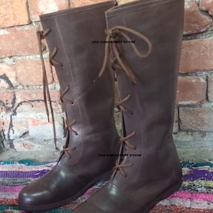 May include: A pair of brown leather knee-high boots with lace-up closures. The boots have a classic design and are suitable for various occasions. The text "USA HANDICRAFT STORE" is visible on the boots. The boots are displayed on a colorful rug.