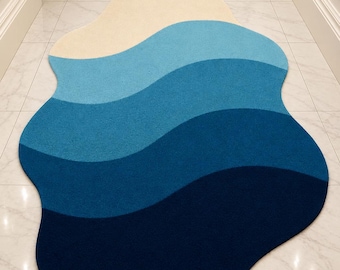 May include: An irregular, organic-shaped area rug featuring a gradient of wavy color bands. The design transitions from a light cream at the top through shades of sky blue, medium blue, and deep teal, ending in a dark navy blue at the base.