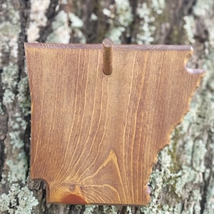 May include: A wooden wall plaque shaped like the state of Arkansas with a medium brown wood grain finish. The plaque features a single small wooden peg protruding from the upper center section, designed for hanging items or display purposes.