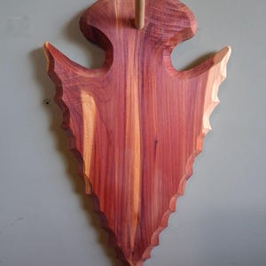 May include: A wall-mounted wooden plaque shaped like a large arrowhead, featuring a prominent reddish-brown wood grain with lighter streaks. A small, cylindrical wooden peg extends vertically from the top center of the arrowhead-shaped board.