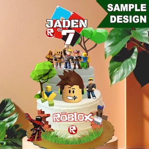 May include: A white frosted cake decorated with Roblox-themed figurines and toppers. The cake features the name "Jaden" and the number "7" on top. The Roblox logo is displayed on the front of the cake, along with a "Sample Design" sign.
