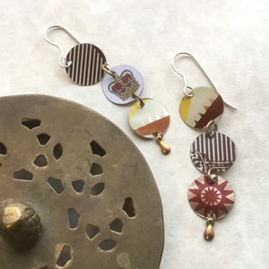 May include: A pair of dangle earrings with multiple round charms. The charms are made of metal and feature a variety of patterns, including stripes, crowns, and geometric designs. The earrings are silver and gold in color.