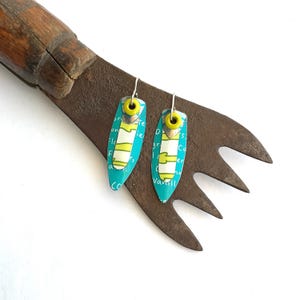 Puede incluir: Un par de pendientes hechos a mano con un fondo turquesa y un diseño amarillo y blanco. Los pendientes son oblongos con un gancho plateado. Los pendientes están sobre una herramienta de metal oxidado.