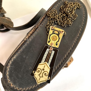 May include: A vintage-style necklace with a geometric pendant featuring black, gold, and cream colors. The pendant hangs from a bronze chain and rests on a black leather surface. The necklace has an Art Deco design.