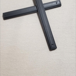 May include: Two black, rectangular objects are arranged in a cross shape. The objects have a textured, ridged surface. The background is a light beige fabric.