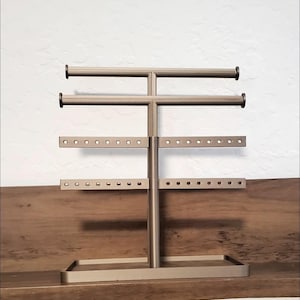 Puede incluir: Un expositor de joyas de color bronce con múltiples barras horizontales y filas de agujeros para pendientes. El soporte tiene una base rectangular y un soporte central vertical. La parte superior tiene dos barras horizontales con tapas negras.