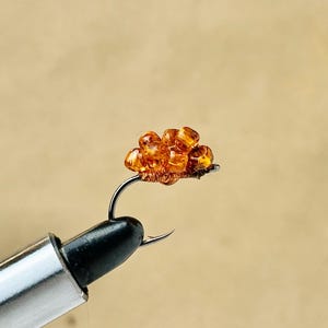 May include: Close-up of a fishing fly with a cluster of orange beads attached to a curved hook. The hook is held by a silver and black tool, set against a neutral background. This fly is designed for catching fish.