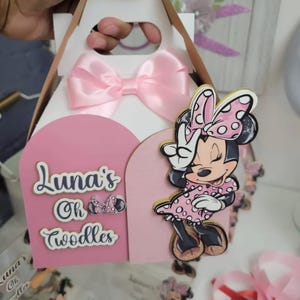 May include: A pink and white party favor box with a Minnie Mouse design. The box has a pink bow, a pink Minnie Mouse cutout, and the text "Luna's Oh Toodles". The box is designed for a child's birthday party.