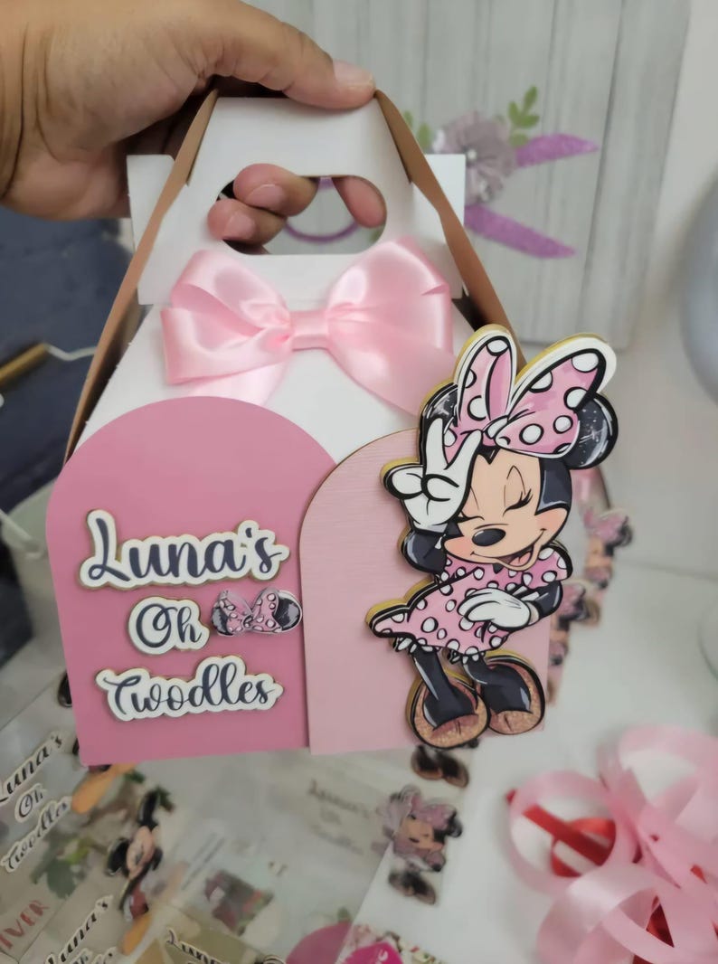 May include: A pink and white party favor box with a Minnie Mouse design. The box has a pink bow, a pink Minnie Mouse cutout, and the text "Luna's Oh Toodles". The box is designed for a child's birthday party.