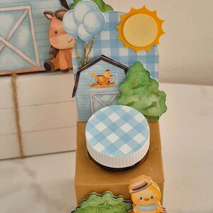 Pode incluir: Uma caixa decorativa com tema de fazenda com tampa xadrez azul e branca. A caixa apresenta recortes de papel de um celeiro, sol, nuvens e um pintinho. A paleta de cores inclui azul, amarelo, marrom e verde.
