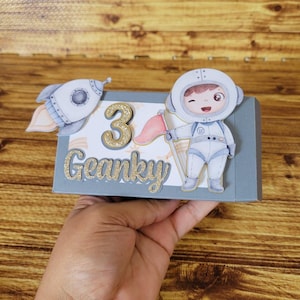May include: A gray box decorated with a rocket ship, the number 3, and the word "Geanky" in gold glitter. An astronaut illustration winks and holds a pink flag. The box is likely for a birthday.