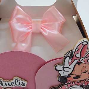 May include: A pink gift box with a large satin bow. The box features a Minnie Mouse cutout and the text "Anelis Oh Goodies". The box is designed for a party or special occasion.