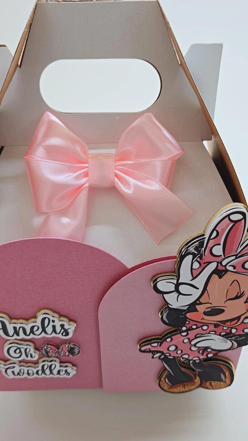 May include: A pink gift box with a large satin bow. The box features a Minnie Mouse cutout and the text "Anelis Oh Goodies". The box is designed for a party or special occasion.