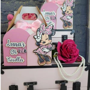 May include: Pink Minnie Mouse-themed party decorations. The display includes a pink suitcase, gift boxes with bows, and Minnie Mouse cutouts. A large pink rose and a pearl necklace add to the festive arrangement. The text "Luna's Oh Toodles" is visible.