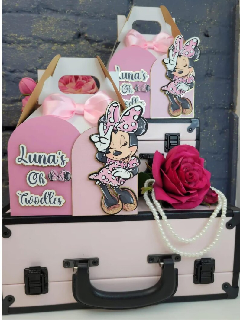 May include: Pink Minnie Mouse-themed party decorations. The display includes a pink suitcase, gift boxes with bows, and Minnie Mouse cutouts. A large pink rose and a pearl necklace add to the festive arrangement. The text "Luna's Oh Toodles" is visible.