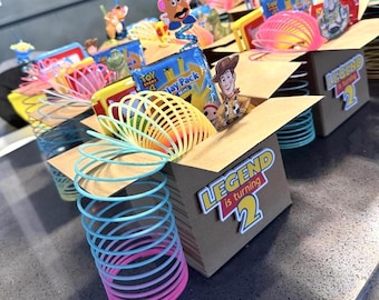 May include: A brown cardboard box filled with Toy Story 4 themed party favors, including a colorful plastic slinky, a Woody cutout, and a Toy Story 4 play pack. The box features a label that reads Legend is turning 2 in blue and yellow text.