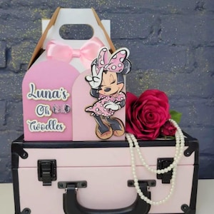 May include: A pink decorative box with a white handle and a pink bow. The box has a Minnie Mouse cutout and the text "Luna's Oh Toodles". A red rose and a pearl necklace are also present.