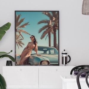 May include: Framed art print featuring a woman in a long dress leaning on a vintage car. The artwork depicts a sunny scene with palm trees and a blue sky. The print is displayed on a white cabinet with a plant and coffee maker.