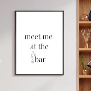 May include: Framed wall art with the text "meet me at the bar" in a black font. The artwork includes a small illustration of a person. The frame is black, and the background is white.